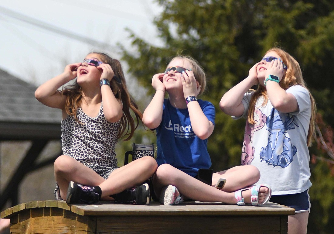 Dunkirk residents, from left, Alyzah Willoughby, 7, Aubrey McClain, 10, and Brionna Walradth, 10, gaze into the sky using their eclipse glasses Monday afternoon from atop the wooden train at Webster Depot Park as they await the moment of totality. The festival drew several hundred visitors to view the eclipse, including one family from Lynchburg, Virginia. (The Commercial Review/Ray Cooney)