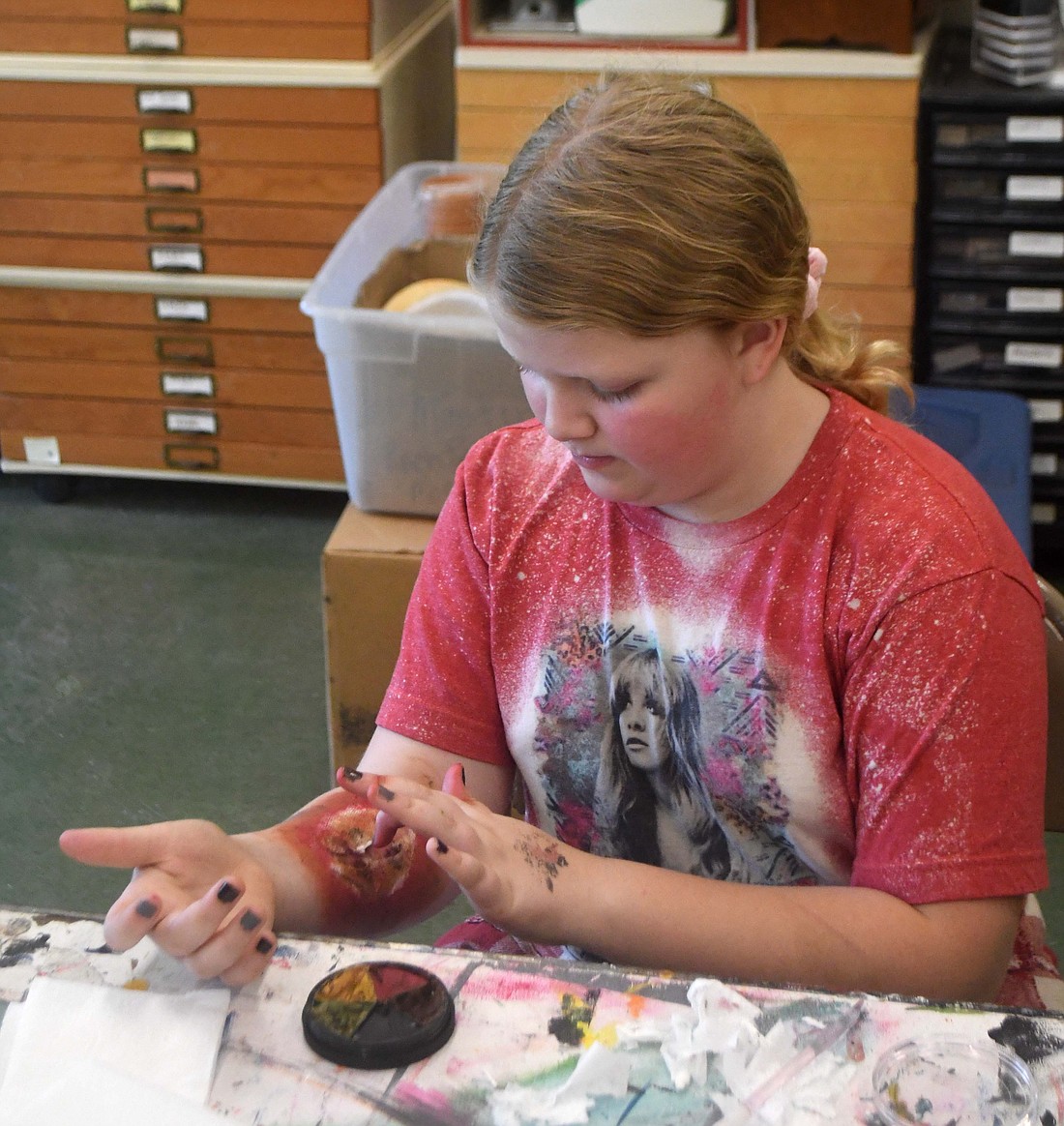 Ten-year-old Kinley Tarter uses her finger to add some detail to her arm during the Zombie FX Makeup Arts in the Park class Thursday morning at the Jay County Campus of Arts Place in Portland. Participants worked with Arts in the Parks instructor Kat Luong to learn how to create zombie-like skin and about special effects makeup. (The Commercial Review/Ray Cooney)