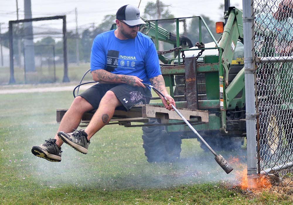 Dunkirk Junior League’s president Deon Jeffers uses a torch to remove weeds and overgrown grass at the bottom of the right foul line fence on Wednesday afternoon. Jeffers, other board members and members of the community have come together the past two months to revive the baseball league for the first time in five years, with the first game on Monday. (The Commercial Review/Andrew Balko)