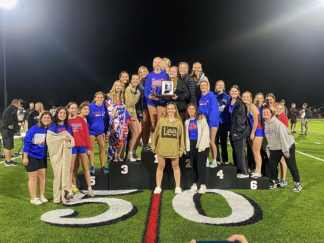 The Jay County High School girls track team poses atop the podium with the Allen County Athletic Conference championship trophy on Friday night at Bluffton. The Patriots only had three victories but a plethora of second and third-place finishes lifted them to victory over Bluffton by 19 points. (Photo provided)